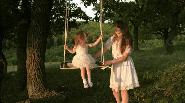 ママは太陽の下で木の下でブランコで彼女の娘を振る。クローズ アップ。母と赤ちゃんは森のオークの枝にロープスイングに乗ります。女の子は笑い、喜ぶ。自然の中で、公園で家族の楽しみ。暖かい夏の日. — ストック写真
