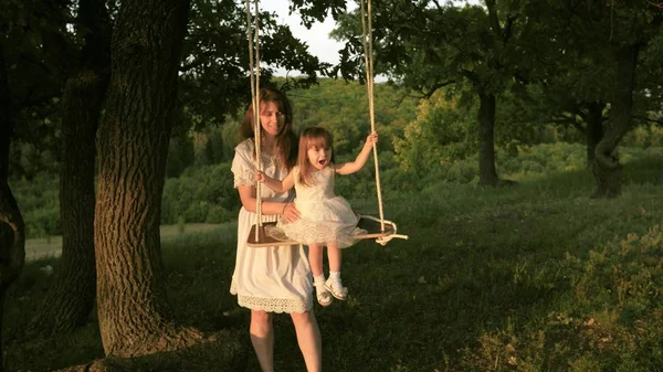 Mamma skakar sin dotter på swing under ett träd i solen. Närbild. mor och baby rida på ett rep Swing på en ek gren i skogen. Flickan skrattar, jublar. Familj kul i parken, i naturen. varma sommardagen. — Stockfoto