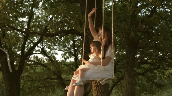 Mamma skakar sin dotter på swing under ett träd i solen. Närbild. mor och baby rida på ett rep Swing på en ek gren i skogen. Flickan skrattar, jublar. Familj kul i parken, i naturen. varma sommardagen. — Stockfoto