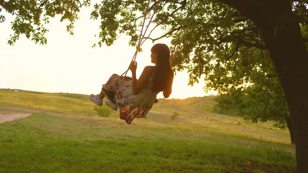 Красивая девушка в платье в парке на качелях мух. Молодая девушка, качающаяся на веревке на дубовой ветке. Девочка-подросток наслаждается полетом на качелях летним вечером в лесу — стоковое фото