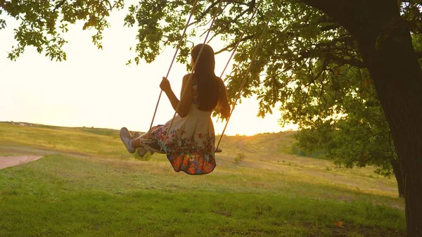 Belle fille dans une robe dans un parc sur une balançoire mouches. jeune fille se balançant sur une balançoire de corde sur une branche de chêne. adolescent fille bénéficie d'un vol sur une balançoire un soir d'été dans la forêt — Photo
