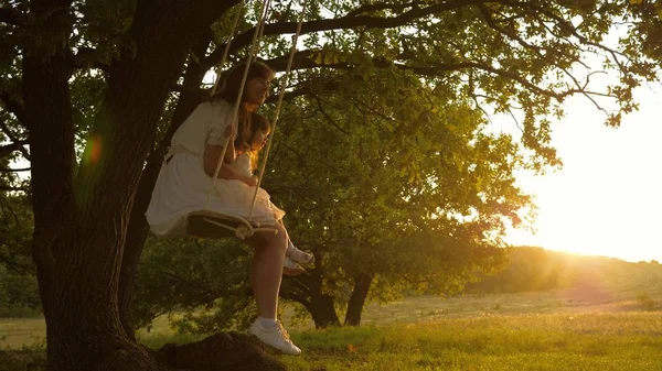 Mamá sacude a su hija en columpio bajo un árbol bajo el sol. madre y el bebé montar en un columpio cuerda en una rama de roble en el bosque. La chica se ríe, se alegra. Diversión familiar en el parque, en la naturaleza. cálido día de verano . —  Fotos de Stock