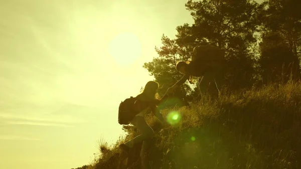 Le ragazze viaggiatrici scalano la montagna, si offrono una mano a vicenda, aiutando a scalare la collina. Famiglia di turisti che viaggiano al tramonto. sorelle con zaini viaggiano scalando la montagna al sole. lavoro di squadra turistico — Foto Stock