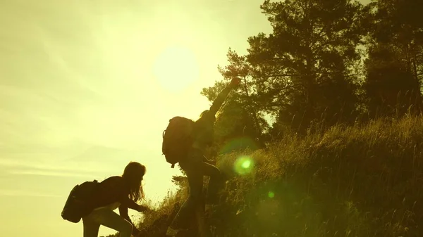 Дівчата-мандрівники піднімаються на гору, пропонують один одному руку допомоги, допомагаючи піднятися на пагорб. Сім'я туристів, які подорожують на заході сонця. сестри з рюкзаками подорожують сходженням на гору на сонці. туристична командна робота — стокове фото