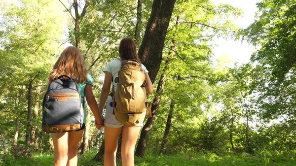 Meninas felizes viajantes com mochilas atravessam o matagal na floresta. crianças turistas viajam no parque de verão. adolescentes à procura de aventura . — Fotografia de Stock