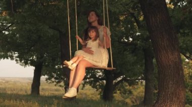 anne ve bebek ormanda bir meşe dalı üzerinde bir ip salıncak binmek. Anne güneşte bir ağacın altında salıncakta kızını sallar. Kız güler, sevinir. Parkta, doğada aile eğlencesi. sıcak yaz günü.