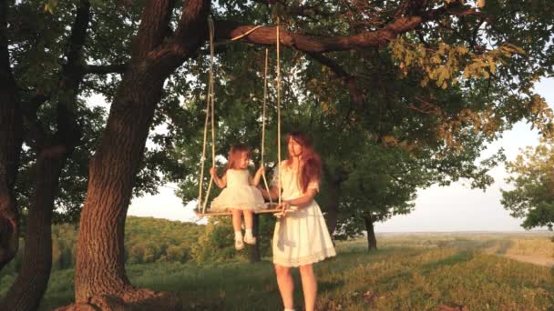 Mamma scuote sua figlia sull'altalena sotto un albero al sole. bambino ride e gioisce. madre e bambino cavalcano su un'altalena di corda su un ramo di quercia nella foresta. Divertimento in famiglia nel parco, nella natura. calda giornata estiva . — Video Stock