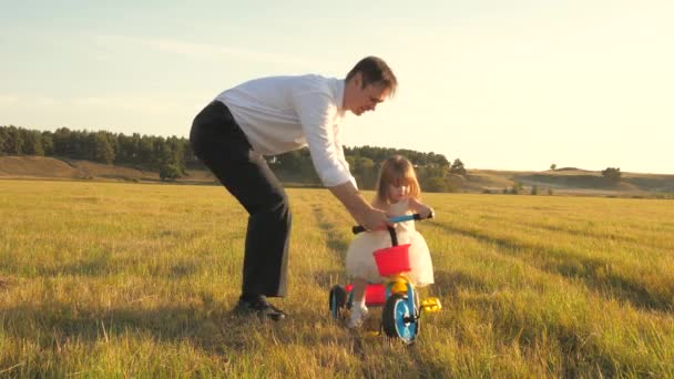 Pai feliz ensina a filha a andar de bicicleta. O pai brinca com uma criança pequena no relvado. O miúdo aprende a andar de bicicleta. pais e filhinha caminha no parque. conceito de família feliz e infância . — Vídeo de Stock