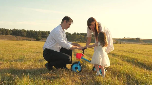 Táta a máma učí dceru jezdit na kole. Matka a otec si hrají s malým dítětem na trávníku. Ten kluk se učí jezdit na kole. rodiče a dceruška se procházejí v parku. koncept šťastné rodiny a dětství — Stock fotografie