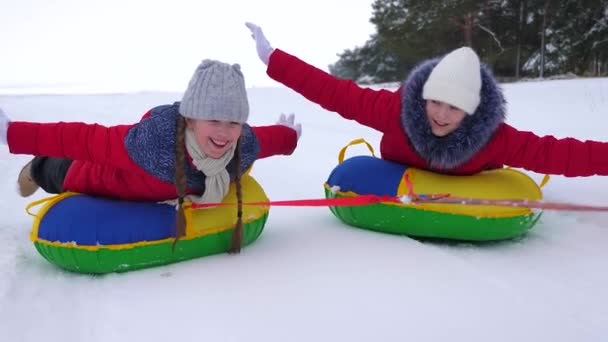 Vacaciones de Navidad para niños al aire libre. Los niños felices se divierten montando platillo de nieve y se ríen en el camino nevado de invierno en el día helado de invierno. Los adolescentes juegan en trineo en el campo de invierno y sonríen. Juegos en fresco — Vídeo de stock