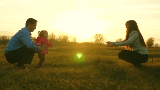Mama en papa spelen met kind op het gras bij zonsondergang. familie geluk concept. baby gaat op gazon van vader naar moeder. kind neemt eerste stappen in park. — Stockvideo