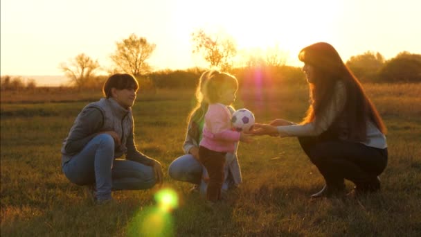 Familie spelen met klein kind door kinderen bal in het park bij zonsondergang. Langzame beweging. Het kind houdt voetbal vast in felle zonnestralen op het veld. gelukkig klein meisje spelen bal met mam en zussen. — Stockvideo