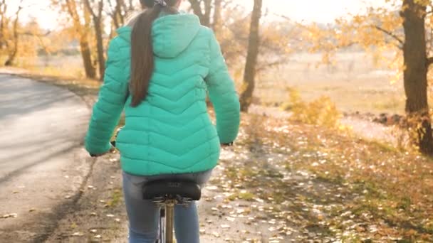 女の子は途中で自転車で旅行します。自転車に乗ってスポーティな十代の若者。緑のジャケットを着た若い女の子が秋の公園へ向かう道で自転車に乗る. — ストック動画