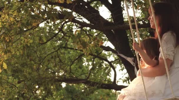 Mamma skakar sin dotter när hon svingar sig under ett träd i solen. närbild. Mamma och baby rida på ett rep swing på en ek gren i skogen. Flickan skrattar, jublar. Familjekul i parken, i naturen. varm sommardag. — Stockvideo