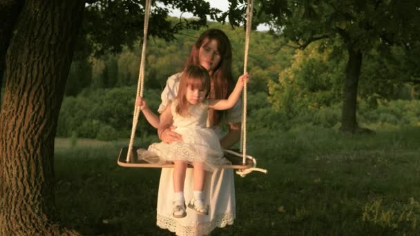 Мама трясет своей дочерью на качелях под деревом на солнце. мать и ребенок катаются на веревке качели на дубовой ветке в лесу. Семейное веселье в парке, на природе. концепция счастливой семьи и детства . — стоковое видео