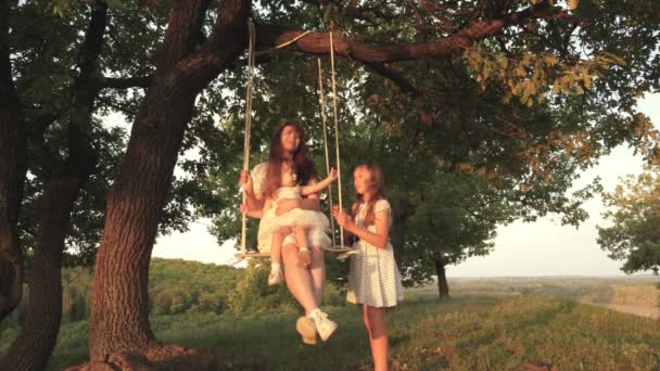 Mamma och baby rida på ett rep swing på en ek gren i skogen. Familjekul i parken, i naturen. Varm sommardag. Mamma skakar sin dotter när hon svingar sig under ett träd i solen. barn skrattar och gläds. — Stockvideo