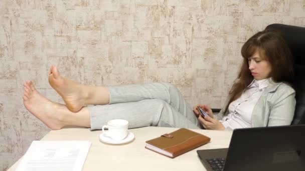 Hermosa mujer de negocios hablando por teléfono, sentado en silla con los pies descalzos en la mesa descansando y bebiendo té, café. chica feliz en el trabajo en la oficina hablando en un teléfono inteligente con una taza de café en la mano . — Vídeo de stock