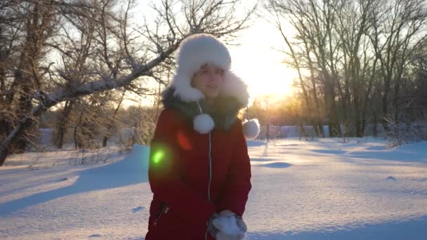 Ragazza felice getta la neve con le mani al tramonto nella foresta. La neve cade e brilla al sole. bambino gioca in inverno nel parco per le vacanze di Natale . — Video Stock
