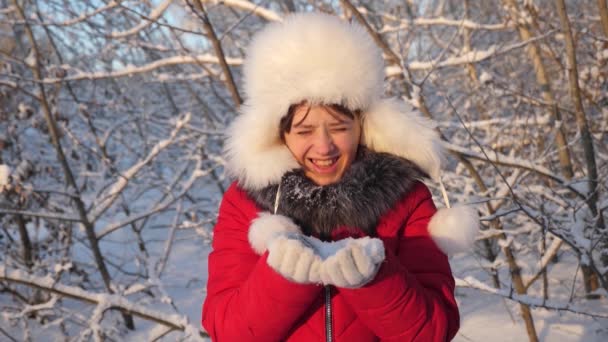 夕暮れ時に雪の結晶を吹いて、冬の公園で笑顔の幸せな女の子。冬の森の中で雪片を手から吹いてる子供はクリスマス休暇で公園で冬に遊びますスローモーション — ストック動画