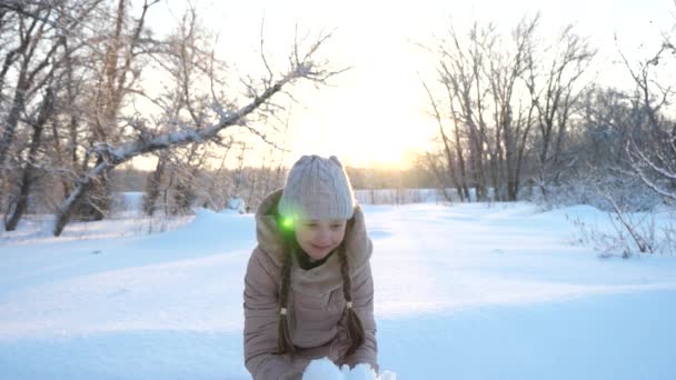 Il bambino gioca in inverno in parco per vacanze di Natale lancia la neve su. bambino gioca in inverno nel parco per le vacanze di Natale. Ragazza felice lancia fiocchi di neve al tramonto e sorride in un parco invernale. Lento. — Video Stock