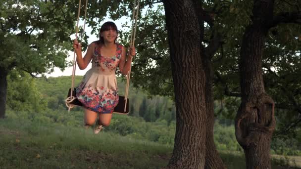 Дитина гойдається на гойдалці в парку на сонці. молода дівчина гойдається на мотузці на дубовій гілці. дівчина-підліток насолоджується польотом на свінгу в літній вечір у лісі. концепція щасливої сім'ї та дитинства . — стокове відео