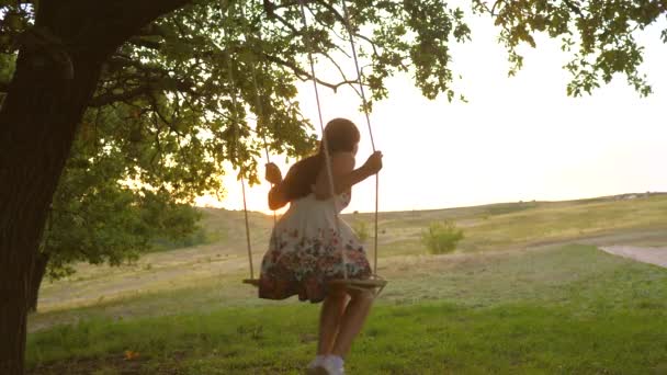 Kind swingend op een schommel in het park in de zon. jong meisje zwaaiend op touw schommel op een eiken tak. tienermeisje geniet van vliegen op swing op zomeravond in het bos. concept van gelukkig gezin en jeugd. — Stockvideo