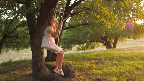 太陽の下で公園でブランコに乗ってる子供。オークの木の枝でロープを振っている少女。10代の女の子は森の夏の夜にスイングで飛行を楽しんでいます。幸せな家族や子供時代の概念は. — ストック動画