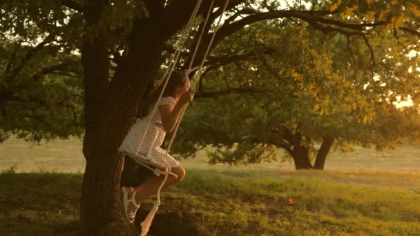 太陽の下で公園でブランコに乗ってる子供。オークの木の枝でロープを振っている少女。10代の女の子は森の夏の夜にスイングで飛行を楽しんでいます。幸せな家族や子供時代の概念は. — ストック動画