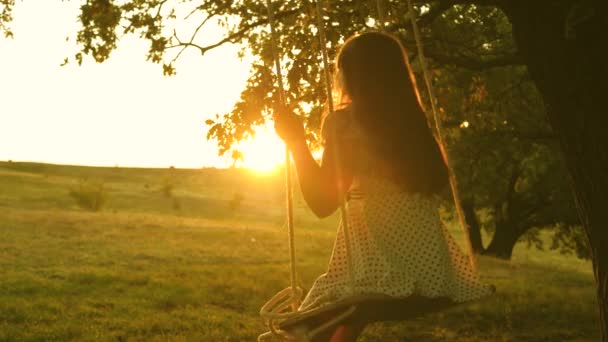 太陽の下で公園でブランコに乗ってる子供。オークの木の枝でロープを振っている少女。10代の女の子は森の夏の夜にスイングで飛行を楽しんでいます。幸せな家族や子供時代の概念は. — ストック動画