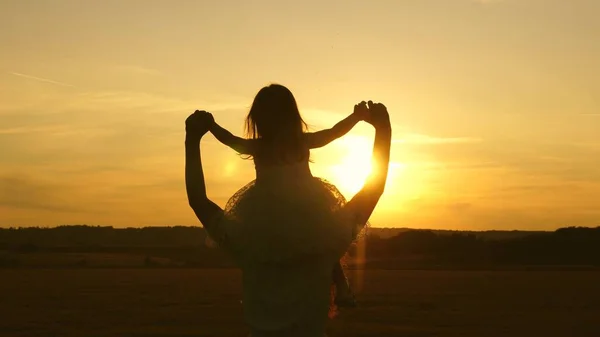 A filha pequena que monta em ombros de mães no por do sol, anda no parque no verão. mãe carrega criança feliz no sol. jovem família com criança no campo. Crianças mãe jogar no prado ao sol . — Fotografia de Stock