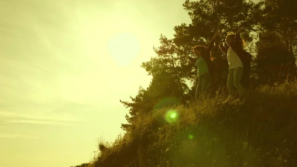 Las chicas viajeras suben a la montaña, saltan y se regocijan en la victoria levantando las manos. Familia de turistas que viajan al atardecer. hermanas con mochilas viajan escalando una montaña al sol. trabajo en equipo turístico —  Fotos de Stock