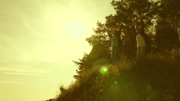 Gezgin kızlar dağa tırmanıyor, atlıyor ve zaferle seviniyor ellerini kaldırıyorlar. Günbatımında seyahat eden turist ailesi. Sırt çantalı kız kardeşler güneşin altında dağa tırmanırlar. turist takım çalışması — Stok fotoğraf