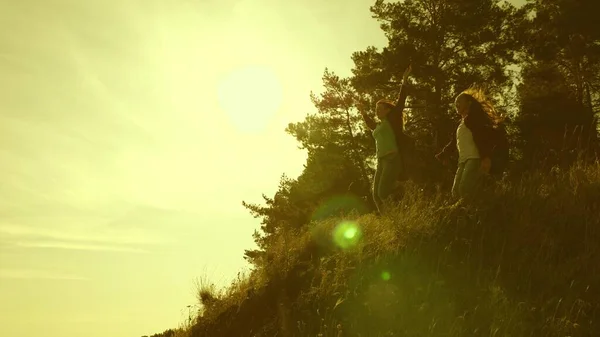 Wandermädchen erklimmen Berge, springen und jubeln über den Sieg, heben die Hände. Touristenfamilie bei Sonnenuntergang unterwegs. Schwestern mit Rucksäcken besteigen einen Berg in der Sonne. Touristische Teamarbeit — Stockfoto