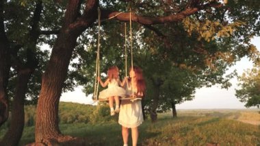 Anne güneşte bir ağacın altında salıncakta kızını sallar. Close. anne ve bebek ormanda bir meşe dalı üzerinde bir ip salıncak binmek. Kız güler, sevinir. Parkta, doğada aile eğlencesi. sıcak yaz günü.