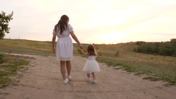 Una pequeña hija con un vestido blanco está caminando con la mano de su madre a lo largo de la carretera. Paseo nocturno de una mujer y un niño. Cuidado materno y afecto. No es una familia completa. Una familia sin padre — Vídeo de stock