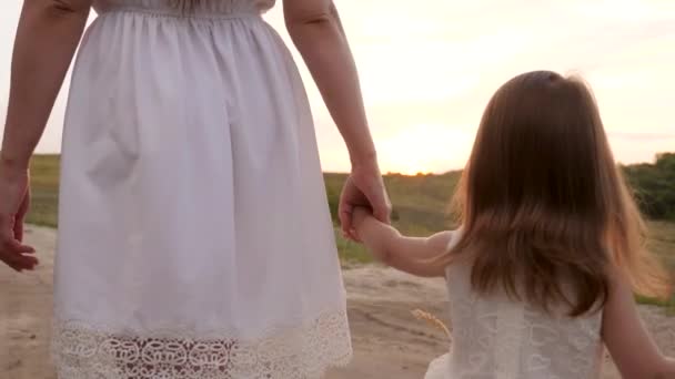 A little daughter in a white dress is walking with her moms hand along the road. Evening walk of a woman and a child. Maternal care and affection. Not a complete family. A family without a father — Stock Video