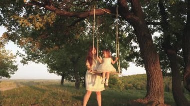 Mutlu anne, neşeli kızını meşe ağacının üzerindeki bir parkta salıncakta sallıyor. Mutlu aile kavramı. Bekar bir anne. Anne bakımı ve çocuk bakımı