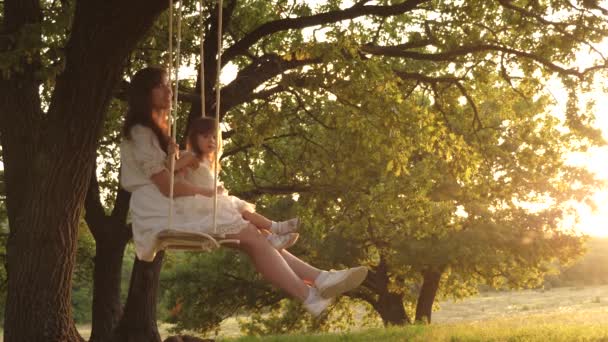 Happy mom rolls a funny baby on a swing in a park on an oak tree at sunset. The concept of a happy family. Single mother. Maternal care and child care. Childhood and flight experience — Stock Video