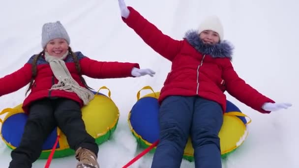 Niños felices adolescentes ruedan en un plato nevado a lo largo de un camino blanco y nevado y se ríen. Chicas alegres van en trineo en invierno y sonríen. Vacaciones de Navidad — Vídeo de stock