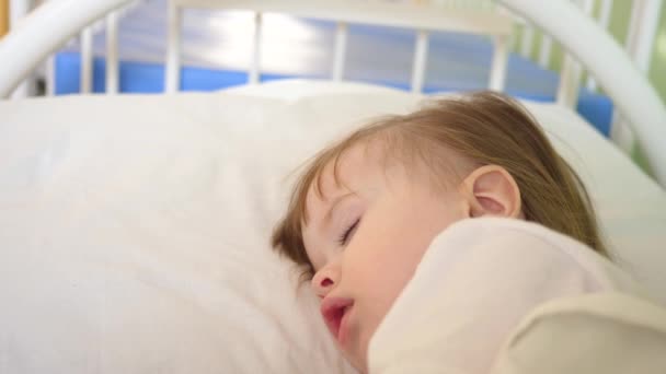 Μικρό παιδί κοιμάται στο νοσοκομείο ward λευκά κλινοσκεπάσματα και μετρά τη θερμοκρασία με θερμόμετρο. Θεραπεία των παιδιών στο νοσοκομείο. Άρρωστο βρέφος βελτιώνει την υγεία του στο νοσοκομείο — Αρχείο Βίντεο