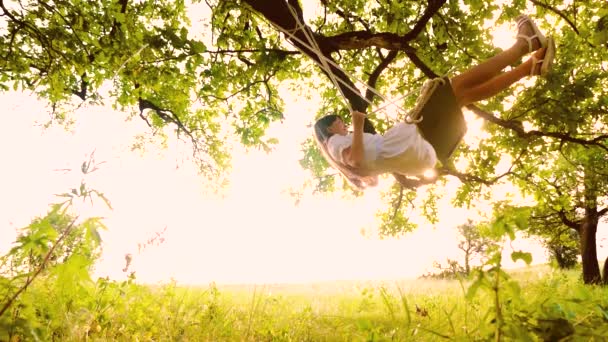 Menina bonita balançando no balanço iluminado pelos raios de sol. corda balançar em galhos de árvores no parque. Movimento lento . — Vídeo de Stock
