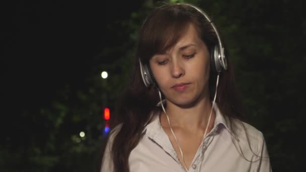 Fille dans le casque comprend chanson dans le téléphone tactile agitant ses cheveux longs et dansant souriant et kayfuya de la musique la nuit dans le parc de la ville. Gros plan — Video