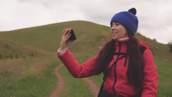 Gratis Giovane ragazza blogger turistica registra video selfie ai piedi delle montagne utilizzando uno smartphone con un bellissimo paesaggio in background. Donna sana allegra viaggia e fotografa la natura per telefono — Foto Stock