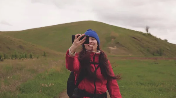 Gratis Giovane ragazza blogger turistica registra video selfie ai piedi delle montagne utilizzando uno smartphone con un bellissimo paesaggio in background. Donna sana allegra viaggia e fotografa la natura per telefono — Foto Stock