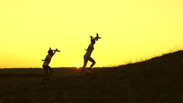 Healthy kids run from mountain with an airplane in hand against backdrop of sun. Dreams of flying. Happy childhood concept. girls play with a toy plane . Silhouette of children playing on plane — Stock Photo, Image