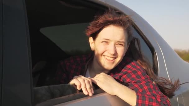 Chica libre con el pelo largo se sienta en el asiento trasero de un coche, mira por la ventana y sonríe al sol. concepto de aventura de coche. feliz joven viaja en coche, se regocija en el viento de la ventana del coche . — Vídeos de Stock