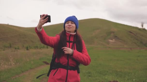 Бесплатная молодая девушка туристический блоггер записывает селфи видео у подножия гор, используя смартфон с красивым пейзажем в фоновом режиме. Здоровая веселая женщина путешествует и фотографирует природу по телефону — стоковое видео