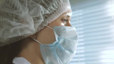 Koruyucu giysili çok yorgun kadın doktor pencereden dışarı bakar, tıbbi maskesini çıkarır, yorgunluktan gözlerini kapatır, temiz hava soluyarak nefes alır. Hastane salgınında doktorların çalışma günü zor geçiyor.