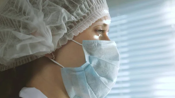 Médico mujer muy cansado en traje protector mira por la ventana, se quita la máscara médica, cerrando los ojos de la fatiga, respira aire fresco. Día laboral difícil del médico en el hospital Epidemia —  Fotos de Stock