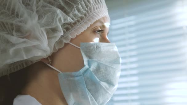 Очень уставшая женщина врач в защитном костюме смотрит в окно, снимает с себя медицинскую маску, закрывает глаза от усталости, дышит свежим воздухом. Трудный рабочий день врача в больнице Эпидемия — стоковое видео